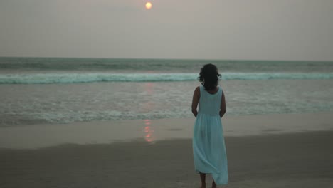 Frau-Im-Weißen-Kleid-Beobachtet-Den-Sonnenuntergang-An-Einem-Ruhigen-Strand,-Der-Sich-Auf-Der-Meeresoberfläche-Spiegelt,-Ein-Ruhiger-Moment