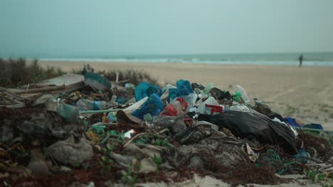 Montón-De-Residuos-Plásticos-En-Una-Playa-Con-Una-Persona-Borrosa-Caminando-Al-Fondo,-Destacando-La-Contaminación.