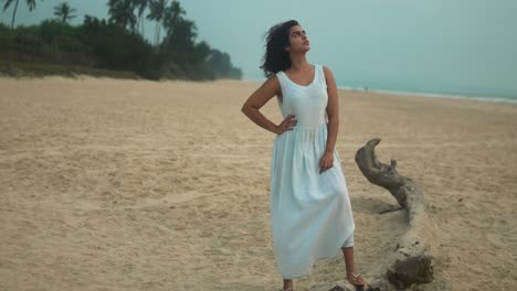 Woman-in-white-dress-posing-thoughtfully-on-a-deserted-beach-at-dusk,-overcast-sky