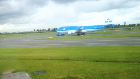 KLM-Airline-Blue-Airplane-pulled-by-truck-tug-on-a-runway-with-green-grass-in-the-front-and-rainy-clouds-in-the-background