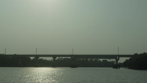 Sunlit-river-shimmering-under-a-silhouette-of-a-large-bridge,-boats-moored-near-lush-greenery