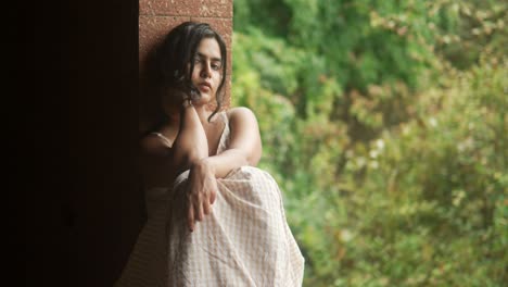 Young-woman-in-a-contemplative-pose-by-a-window,-looking-thoughtful,-with-lush-greenery-in-the-background,-soft-focus