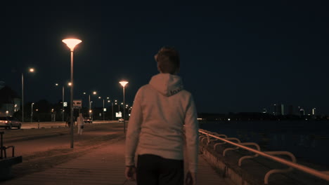 Follow-back-and-a-panoramic-shot-of-Young-man-start-walking-night-time-in-a-modern-city,-urban-landscape-with-the-illuminated-road,-well-lit-street-waterfront