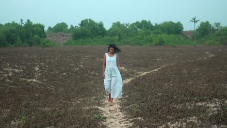 Mujer-Vestida-De-Blanco-Caminando-Por-Un-Camino-De-Tierra-A-Través-De-Un-Campo-árido,-Transmitiendo-Una-Sensación-De-Soledad-Y-Reflexión.