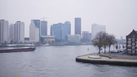 Großes-Frachtschiff,-Das-Durch-Die-Stadt-Rotterdam-Fährt,-Mit-Den-Wolkenkratzern-Des-Finanzviertels-Im-Hintergrund-Und-Langsamem-Schwenk-Auf-Einen-Boulevard-Und-Ein-Wohnviertel-Im-Vordergrund