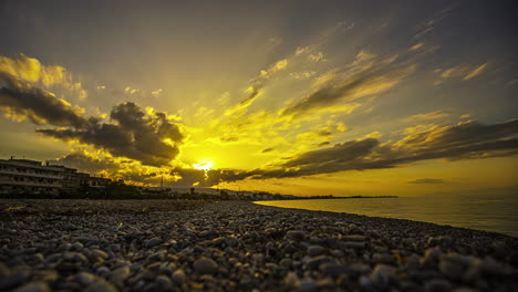 Sun-sets-below-clouds-shooting-light-rays-out-across-pebble-beach,-low-angle