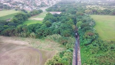 drone-flying-over-a-watery-sewerage-canal-surrounded-by-bush-and-sports-fields-abandon-shacks