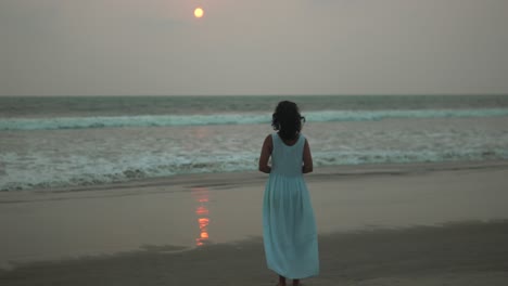 Woman-in-blue-dress-walks-alone-on-beach-at-sunset,-reflective-mood