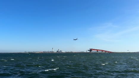 Wide-view-of-an-airplane-flying-over-a-large-body-of-water-near-a-red-bridge-and-industrial-cityscape