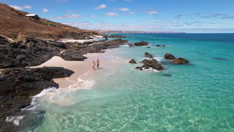 Ein-Paar-Geht-Hand-In-Hand-An-Einem-Weißen-Sandstrand-In-Das-Strahlend-Blaue,-Kristallklare-Meer-Und-Beginnt-Zu-Schwimmen-Und-Zu-Spielen,-Während-Sonst-Niemand-In-Sicht-Ist