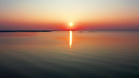 Aerial-Drone-View-of-Beautiful-Ocean-Sunrise-with-Bright-Glowing-Sun-Casting-Colorful-Red,-Orange,-Purple-and-Yellow-Reflections-Over-Peaceful-Rippling-Waves-of-the-Sea-with-Gorgeous-Sky-and-Water