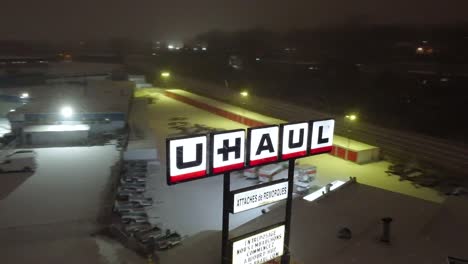 Snowy-night-at-a-U-Haul-rental-facility-in-Montreal,-glowing-signs-light-up-the-tranquil-scene,-aerial-view
