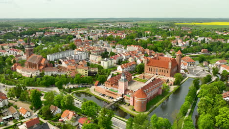 Eine-Weite-Luftaufnahme-Von-Lidzbark-Warmiński,-Die-Das-Historische-Schloss,-Den-Fluss,-Die-Umliegenden-Gebäude,-Grüne-Bäume-Und-Eine-Weite-Sicht-Auf-Die-Stadt-Und-Die-Landschaft-Einfängt