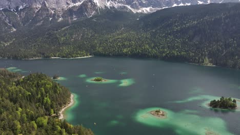 Eibsee's-clear-turquoise-waters,-small-islands,-and-dense-forests-with-a-backdrop-of-snow-capped-mountains-in-Grainau,-Germany