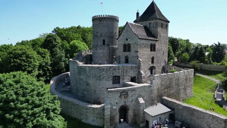 Medieval-castle-with-a-turret,-white-stone-walls,-and-courtyard-during-a-beautiful-summer-day-surrounded-by-lush-greenery,-under-a-clear-blue-sky