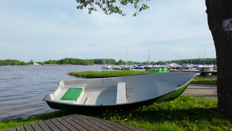 A-small-white-rowboat-resting-on-the-shore-of-Ukiel-Lake-in-Olsztyn,-with-a-marina-and-sailboats-in-the-background