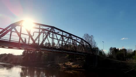 Drone-Vuela-Sobre-El-Río-Contrastado-Puente-Metálico-Cruzando-Agua-Sol-Horizonte