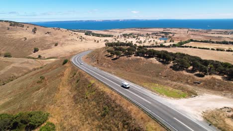 Drone-footage-flying-backwards-as-a-camper-van-and-car-drive-along-a-road-surrounded-by-stunning-nature-and-landscape-in-the-fleurieu-peninsula,-South-Australia