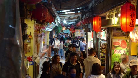 Large-crowd-of-people-wanders-through-a-bustling,-narrow-lane-flanked-by-food-stalls,-souvenir-boutiques,-and-quaint-gift-shops-in-Jiufen-Old-Street,-a-charming-mountain-village-in-Taiwan