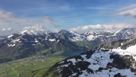 Slow-Aerial-Dolly-Towards-Snow-Covered-Mountain-Peaks-with-Village-and-Blue-Lake-in-Foreground