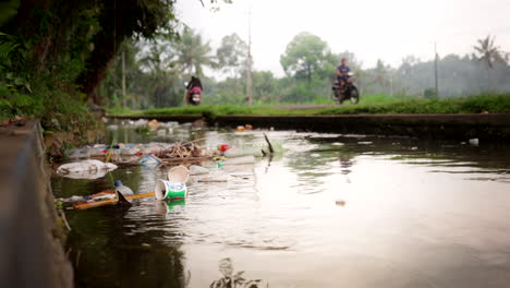 Einheimische-Auf-Motorrollern-Passieren-Stark-Verschmutzte-Wasserstraße-Mit-Schwimmenden-Trümmern,-Bali