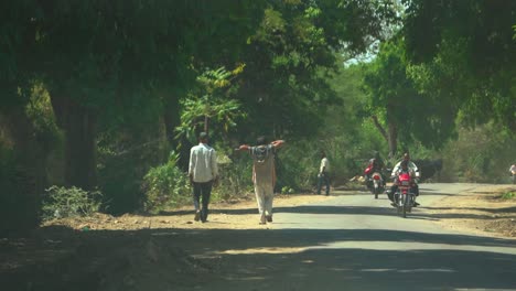 Villagers-and-motorbikes-passing-down-a-village-road-covered-with-lush-green-trees-during-a-summer-afternoon-in-rural-India