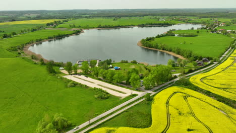 Eine-Luftaufnahme-Des-Wielochowskie-Sees-Und-Seiner-Umgebung,-Einschließlich-Grüner-Felder,-Gelber-Rapsfelder-Und-Einer-Straße,-Mit-Panoramablick-Auf-Die-Landschaft