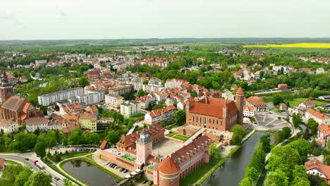 A-broad-aerial-view-of-Lidzbark-Warmiński-focusing-on-the-significant-historical-building-near-a-water-body,-surrounded-by-modern-and-historical-buildings-and-lush-greenery