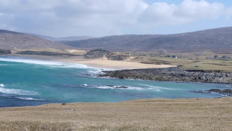 Pan-of-stunning-turquoise-blue-ocean-with-waves-rolling-in-towards-coastal-rural-countryside-landscape-on-the-isle-of-Lewis-and-Harris,-Outer-Hebrides,-Western-Scotland-UK
