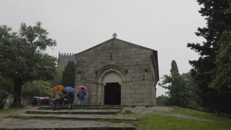 Los-Turistas-Sostienen-Paraguas-Mientras-Caminan-En-Una-Visita-Guiada-En-Un-Día-Lluvioso-Pasando-Por-La-Capilla-De-Piedra.