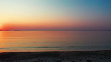 Vista-Panorámica-Derecha-Del-Hermoso-Amanecer-En-La-Playa-Con-Un-Sol-Brillante-Que-Proyecta-Un-Colorido-Reflejo-Rojo,-Naranja,-Morado-Y-Amarillo-Sobre-Las-Tranquilas-Olas-Del-Mar-Con-Un-Hermoso-Cielo-Y-Agua-Del-Océano