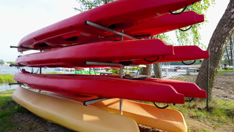 Una-Pila-De-Kayaks-Rojos-Almacenados-En-Estantes-Metálicos-Junto-Al-Lago,-Con-árboles-Y-Agua-Visibles-Al-Fondo