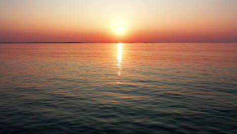Luftaufnahme-Eines-Wunderschönen-Sonnenuntergangs-Am-Meer-Mit-Einer-Hellen,-Leuchtenden-Sonne,-Die-Farbenfrohe-Rote,-Orange,-Violette-Und-Gelbe-Reflexe-über-Friedliche,-Plätschernde-Wellen-Des-Meeres-Mit-Wunderschönem-Himmel-Und-Wasser-Wirft