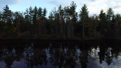 Left-to-right-trucking-shot-of-a-dark-and-moody-tree-filled-shoreline-and-its-reflection-in-a-calm-lake-in-Maine-during-the-Fall