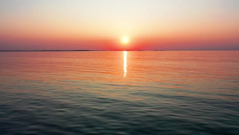 Luftaufnahme-Eines-Wunderschönen-Sonnenuntergangs-Am-Meer-Mit-Einer-Hellen,-Leuchtenden-Sonne,-Die-Farbenfrohe-Rote,-Orange,-Violette-Und-Gelbe-Reflexe-über-Friedliche,-Plätschernde-Wellen-Des-Meeres-Mit-Herrlichem-Himmel-Und-Wasser-Wirft