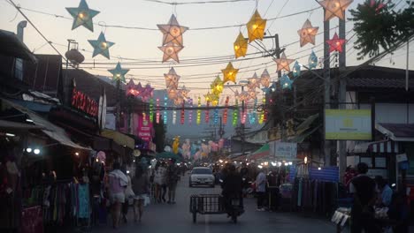 Several-tourists-in-summer-clothes-shop-across-the-street-lined-with-store-stalls-on-a-warm-summer-evening-in-the-city-of-Pai-in-Thailand