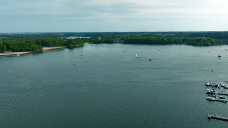 Una-Vista-Aérea-Del-Lago-Ukiel-En-Olsztyn,-Con-Barcos-Y-Yates-En-El-Agua,-Rodeado-De-Bosques-Verdes-Y-Una-Costa-Lejana