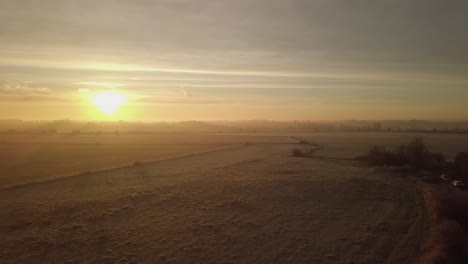 Drone-flies-across-frosty-fields-towards-beautiful-sunrise,-with-the-camera-slightly-panning-up