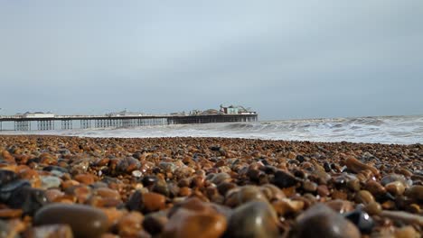 Low-Angle-View-of-Brighton-Pier-with-Pebbles-in-Foreground-and-Waves-Crashing-in-Slow-Motion