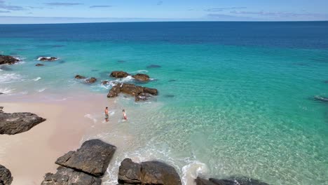 Ein-Paar-Hält-Sich-An-Den-Händen-Und-Geht-Von-Einem-Weißen-Sandstrand-In-Das-Strahlend-Blaue,-Kristallklare-Meerwasser-Hinaus