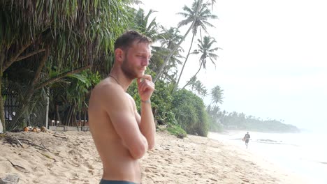 Attractive-Shirtless-Man-Standing-on-Tropical-Sandy-Beach-Looking-Out-Over-the-Water-Evaluating-the-Surfing-Conditions