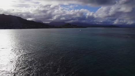 drone-footage-over-Lanakai-Beach-the-island-of-Oahu-Hawaii-with-ocean-in-the-foreground-and-mountainous-islands-on-the-horizon-the-sky-if-filled-with-clouds-preparing-to-rain-a-lone-sailboat