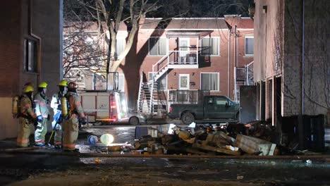 Bomberos-De-Noche-Evaluando-Las-Consecuencias-De-Un-Incendio-En-Un-Edificio-En-Montreal,-Escombros-En-Primer-Plano,-Luces-De-Vehículos-De-Emergencia