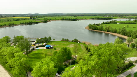 An-aerial-view-of-Wielochowskie-Lake-surrounded-by-green-fields,-trees,-and-a-small-recreational-area-with-a-few-buildings