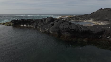 Rocky-coastline-of-Mosteiros,-Sao-Miguel-with-tranquil-tidal-pool-and-rugged-landscape,-sunny-day,-wide-shot