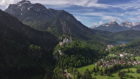 Neuschwanstein-castle-nestled-in-the-bavarian-alps,-surrounded-by-lush-greenery-and-a-small-village,-aerial-view