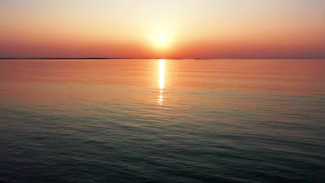 Luftaufnahme-Eines-Wunderschönen-Sonnenuntergangs-Am-Meer-Mit-Einer-Hellen,-Leuchtenden-Sonne,-Die-Farbenfrohe-Rote,-Orange,-Violette-Und-Gelbe-Reflexe-über-Friedliche,-Plätschernde-Wellen-Des-Meeres-Mit-Wunderschönem-Himmel-Und-Wasser-Wirft