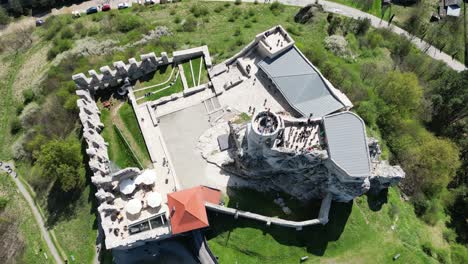 Medieval-Rabsztyn-castle-with-a-tower,-walls,-umbrellas-and-courtyard-during-a-beautiful-summer-day-surrounded-by-lush-greenery,-grass,-and-trees