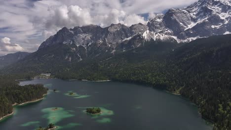 Panorámica-Aérea-Alrededor-De-Un-Gran-Lago-Azul-Situado-En-Un-Valle-De-Montaña-Con-Un-Bosque-Verde-Y-Un-Pico-Cubierto-De-Nieve-En-El-Fondo.