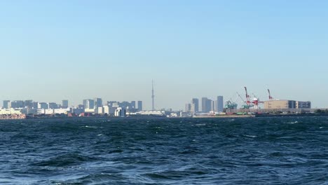 City-skyline-and-industrial-harbor-viewed-across-choppy-blue-sea-waters,-clear-sky